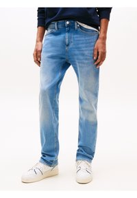 Homme portant un jean slim bleu clair et des baskets blanches, debout, les mains relâchées le long du corps, sur un fond uni.