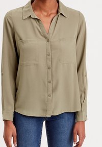 Blouse vert olive avec col, manches longues et deux poches poitrine. Le tissu est lisse avec un léger brillant, doté d'une fermeture à boutons sur le devant.