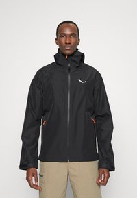 Veste imperméable noire avec un col haut, fermeture éclair, accents orange et un logo sur la poitrine ; dispose de poches des deux côtés.