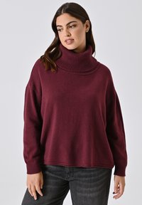 Weinroter Rollkragenpullover mit einem gerippten Kragen, lockerer Passform, weichem Material und langen Ärmeln, kombiniert mit dunklen Jeans.