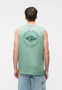 Νέος άνδρας με κοντά καστανά μαλλιά, που φοράει πράσινη αμάνικη μπλούζα Rip Curl Surf Co και ανοιχτόχρωμο τζιν σορτς, γυρισμένος με την πλάτη σε λευκό φόντο.