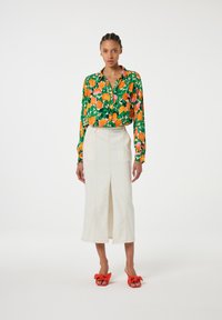 Bloemenblouse met een oranje en groen patroon, hoge witte rok met een split, en rode strik sandalen. Soepele texturen en een getailleerde pasvorm.