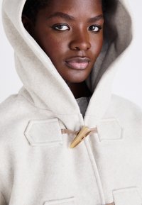 Manteau en mélange de laine blanc avec une capuche, présentant une fermeture à bascule, un tissu texturé et un design minimaliste. Les accents incluent des patchs cousus.