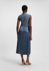Modrá bezrukávová šaty s texturovaným vrškem a sukni podobnou saténu, sahající nad kotníky. Pohled zezadu ukazuje hladkou látku a přiléhavý pas.