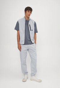 Lysegrå genservest med glidelås og en kontrasterende mørk lomme, kombinert med en mørkegrå T-skjorte og matchende lysegrå joggebukser. Hvite joggesko.