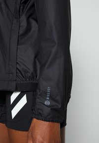 Acercamiento del brazo de una persona que lleva una chaqueta cortavientos negra con el texto "WIND.RDY", combinada con pantalones cortos negros que tienen rayas diagonales blancas.