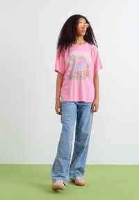T-shirt rose avec un design graphique représentant un arc-en-ciel et un soleil, associé à un jean évasé bleu clair et des chaussures roses. Cheveux bouclés et lunettes de soleil.