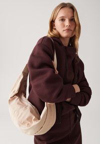 Borsa hobo beige con una texture liscia e una silhouette curva, dotata di una chiusura con zip e indossata sopra un outfit color maroon con colletto alto.