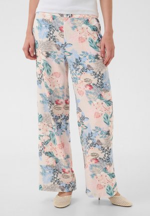 Femme portant un pantalon ample à motif floral dans des couleurs pastel avec des feuilles et fleurs vertes, bleues, roses et rouges, associé à des chaussures tressées beiges.