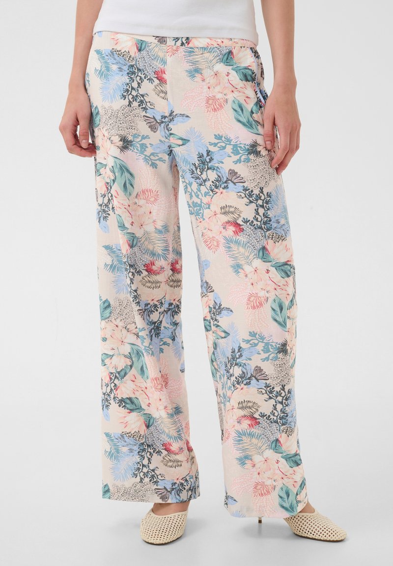 Femme portant un pantalon ample à motif floral dans des couleurs pastel avec des feuilles et fleurs vertes, bleues, roses et rouges, associé à des chaussures tressées beiges.