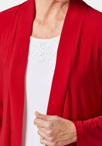 Rode cardigan gemaakt van zachte, gladde stof met een gedrapeerde voorkant, gelaagd over een witte top met een kant ontwerp rond de halslijn.