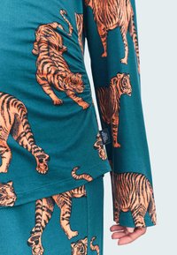 Maglietta a maniche lunghe in tessuto teal decorata con stampe di tigre arancioni. Presenta arricciature sul lato e un'etichetta sull'orlo. Texture liscia.