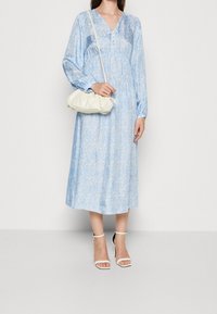 Vestido midi azul de flores con mangas largas y escote en V. Un par de tacones blancos con tiras y un bolso de hombro color crema. Textura suave.