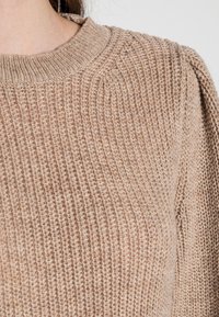 Beżowy sweter z dzianiny o prążkowanej powierzchni, z okrągłym dekoltem i lekko bufiastymi ramionami. Prosty i miękki materiał.