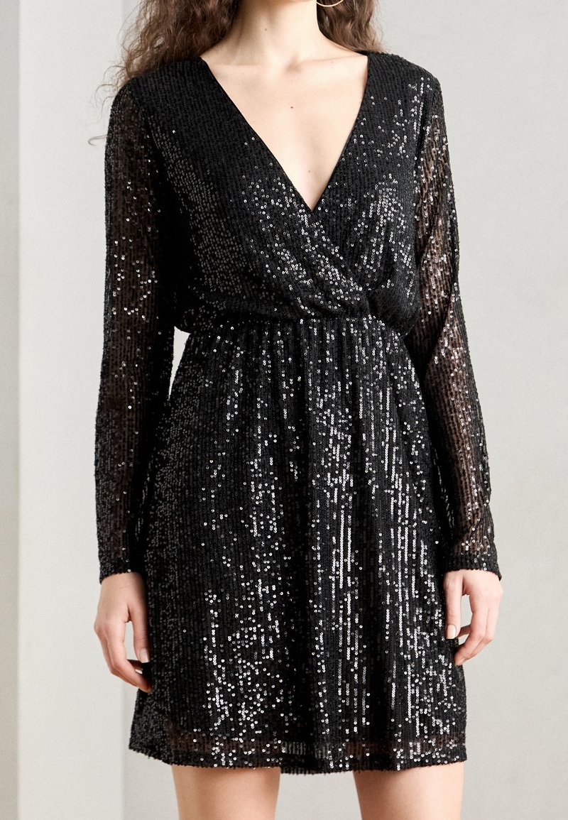 Femme portant une robe noire à manches longues ornée de sequins, avec un décolleté en V profond et une taille froncée.