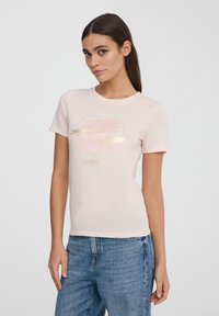 T-shirt di cotone rosa chiaro con maniche corte, caratterizzato da una grafica di cuori oro e pastello e testo sottostante. Texture liscia, design aderente.