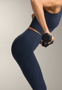 Tricou sportiv fără mâneci, de culoare albastru închis, și colanți cu talie înaltă, având o textură fină și cusături proeminente, ținând în mână o halteră hexagonală.
