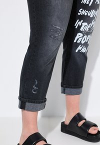 Jeans de mezclilla negra con detalles desgastados, puños enrollados y texto gráfico blanco; combinados con sandalias de plataforma negras.