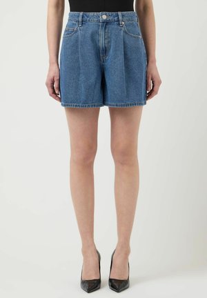 Kvinne iført høytlivete, folderede denimshorts kombinert med svarte spisse hæler, stående foran en enkel lys bakgrunn.