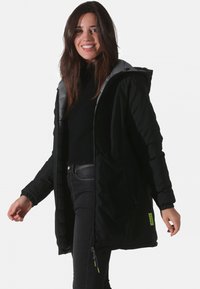 Manteau long noir avec capuche, doté d'une doublure grise, d'une fermeture éclair à l'avant et de poignets élastiques. Comprend une étiquette de marque visible en vert fluo.
