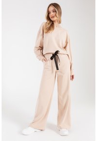 Beżowy zestaw dresowy składający się z luźnej bluzki z opadającymi ramionami oraz spodni z elastycznym pasem i czarną kokardą, w połączeniu z białymi sneakersami.