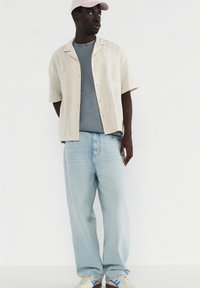 Herrenoutfit mit einem lockeren beigen Kurzarmhemd, grauem T-Shirt, hellblauen Jeans und weißen Sneakers mit blauen Akzenten.