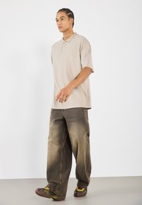 Beige polo met korte mouwen, gecombineerd met loszittende, vervaagde bruine jeans en gemusterde sneakers in tinten groen en rood.