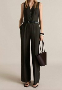 Dunkelgrüner, gerippter Jumpsuit mit tiefem V-Ausschnitt und Knöpfen, an der Taille mit einem schwarzen Gürtel gerafft. Kombiniert mit einer dunklen Handtasche und Sandalen.