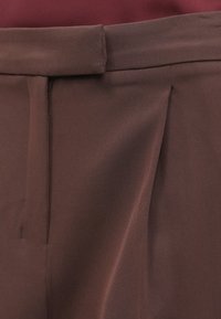 Közelkép sötétbarna nadrágról, derékrésszel, cipzáros nyitással és plissézett elülső részlettel.