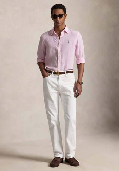 Mees kannab päikeseprille, heleroosat pika varrukaga särki, valgeid pükse, pruuni vööd, pruune kingi ja käekella, seisab ühe käega taskus.