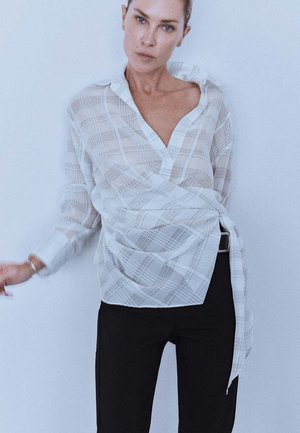 Mujer con una blusa cruzada de cuadros blancos holgada y pantalones negros, de pie contra un fondo claro y liso, mirando hacia adelante.
