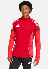 Maglia sportiva rossa a maniche lunghe con zip a mezze, accenti bianchi e tre strisce bianche sulle spalle. Realizzata in tessuto traspirante.
