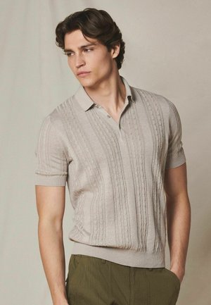 Młody mężczyzna ubrany w beżową krótkorękawową dzianinową koszulkę polo z warkoczowymi wzorami i oliwkowo zielone spodnie, stojący na jednolitym tle.