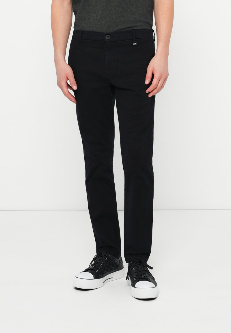 Čierne bavlnené nohavice slim fit so zapínaním na gombík, pútkami na opasok a bočnými vreckami. Nosí sa s čiernymi textúrovanými teniskami.