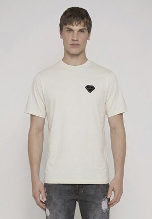 Ung mand, der vender fremad, iført en ensfarvet lys beige kortærmet t-shirt med en lille sort lap på brystet og slidte mørke jeans.
