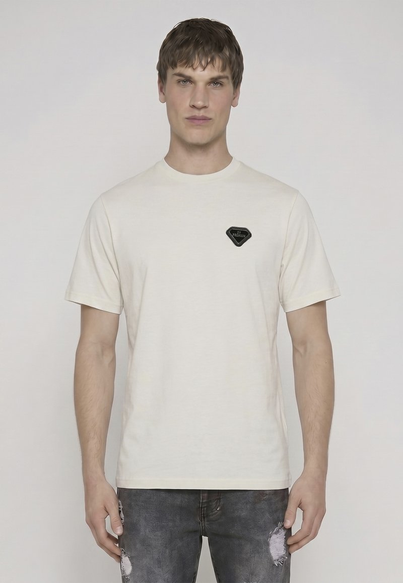Jeune homme face à l'avant portant un t-shirt uni beige clair à manches courtes avec un petit patch noir sur la poitrine et un jean sombre usé.