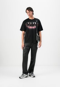Young man wearing a black t-shirt with a large shark teeth graphic, black cargo pants, silver-white sneakers, and a black crossbody bag.