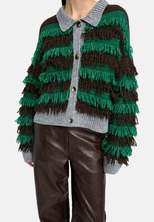 Szary dzianinowy kardigan z zielono-brązowymi teksturowanymi frędzlami i drewnianymi guzikami, noszony z ciemnobrązowymi skórzanymi spodniami.