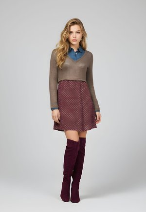 Mujer joven de pie, con un suéter corto marrón sobre una camisa azul, falda burdeos con lunares y botas altas a juego, sobre un fondo gris.