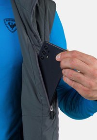 Graue Steppweste mit einem Reißverschluss und einer Tasche für ein schwarzes Smartphone. Die Weste wird über einem blauen Langarmhemd getragen, das eine glatte Textur aufweist.