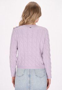 Maglione in maglia lavanda con motivo a trecce, caratterizzato da un scollo rotondo e dettagli con bottoni sulla spalla. Abbinato a una gonna di jeans.