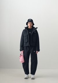 Black windbreaker jacket with two front pockets, paired with matching pants. Accessories include a black bucket hat and a small pink bag.