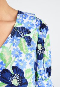 Bloes met bloemen in blauw en groen op een witte achtergrond, met een gekartelde kraag en textuurpofmouwen.