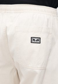 Μπεζ βαμβακερό παντελόνι με ελαστική μέση, που διαθέτει πίσω τσέπη και μαύρο κεντητό λογότυπο με λευκό περίγραμμα.