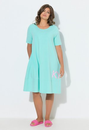 Mujer de pie con un vestido turquesa hasta la rodilla, con mangas cortas y zapatillas rosas, sobre un fondo blanco.