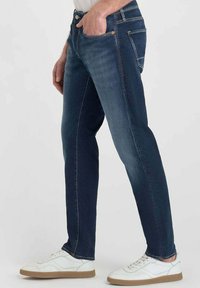 Vaqueros de denim azul oscuro con un corte ajustado, que cuentan con cinco bolsillos, costuras visibles y se llevan con zapatillas blancas. Textura suave.