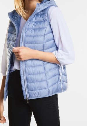 Kvinde iført lyseblå vatteret ærmeløs vest over hvid bluse med lange ærmer og sorte bukser, med vestens lynlås delvist åben.