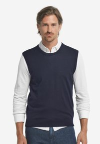 Marinblå ärmlös tröja i mjukt stickat material. Har en rund krage och är lager på lager över en vit långärmad skjorta.