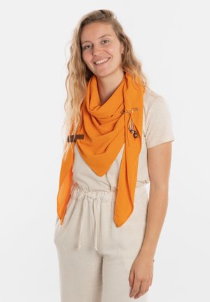 Een oranje textuur sjaal in een driehoekige vorm gedrapeerd met een decoratieve speld en kwastdetail, gedragen over een lichtgekleurde top en broek.