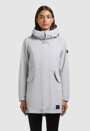 Woman facing forward wearing a light gray hooded coat with flap pockets and a black patch on the left sleeve, against a plain background.